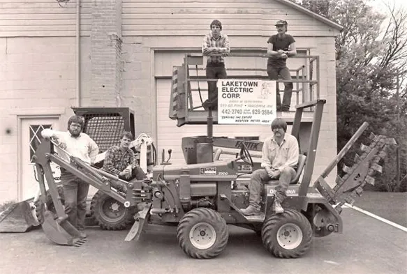 Black and white photo of six early Laketown Electric crew members posed on a trencher and a lift that carries a Laketown Electric Corp. sign